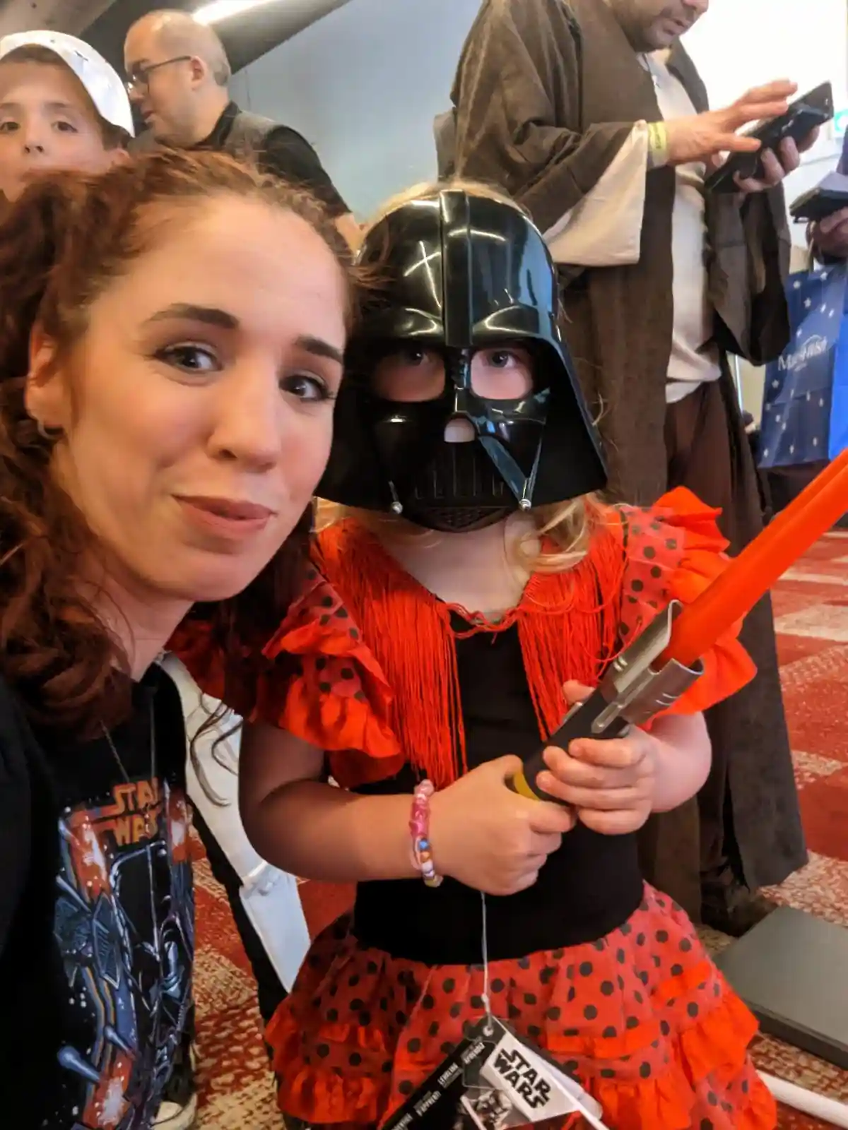 A woman and a young girl in a Star Wars-themed setting pose for a photo. The girl wears a Darth Vader helmet and holds an…