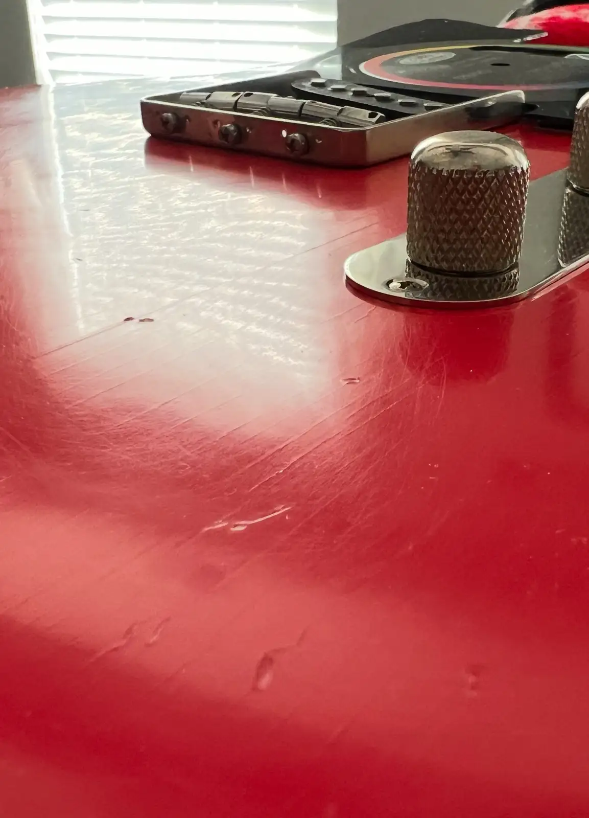 A close-up photo of a red guitar body with visible scratches and dents, featuring a chrome tuning peg in the foreground and…