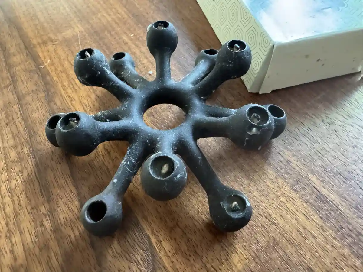 a black, star-shaped candle holder with multiple arms and sockets sits on a wooden surface, visibly dirty with dust and…