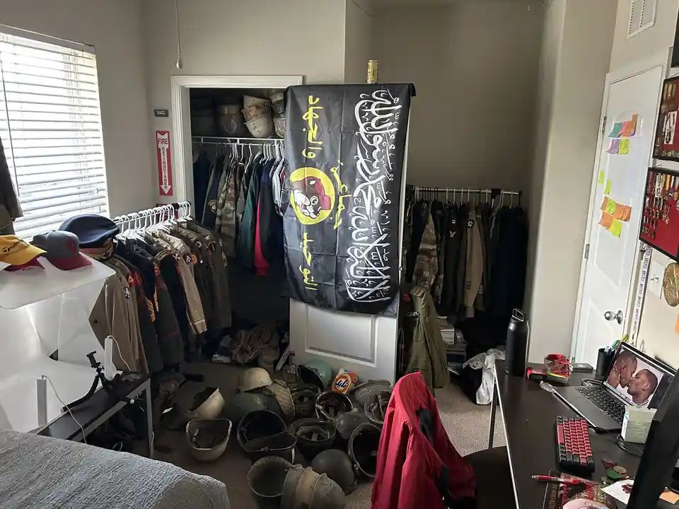 A cluttered bedroom filled with military uniforms, helmets, and gear, centered around a black flag with Arabic script and a…