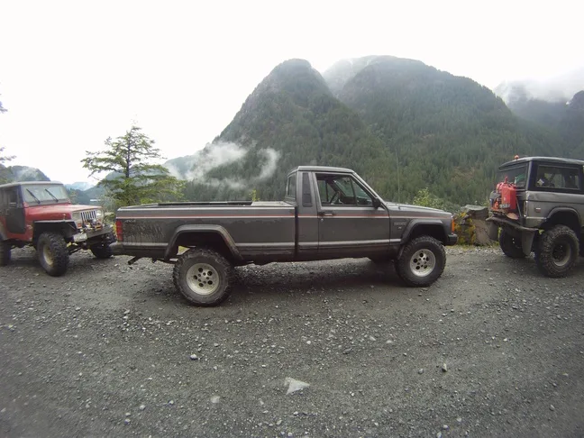 Three rugged off-road vehicles—a red Jeep, a gray pickup truck, and a black SUV—are parked on a gravel road with misty…