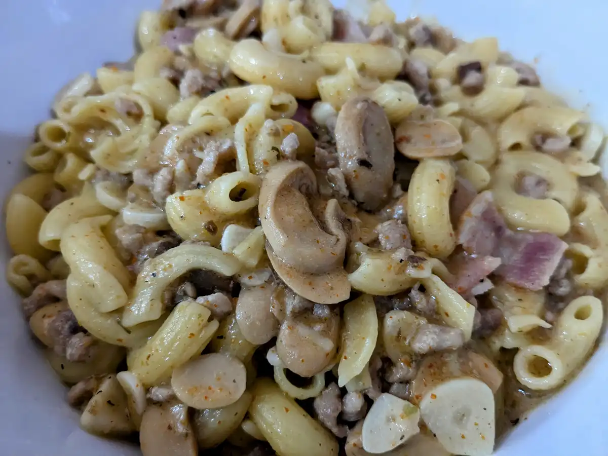 A close-up photo of a white bowl filled with elbow macaroni mixed with ground meat, sliced mushrooms, and garlic cloves in a…
