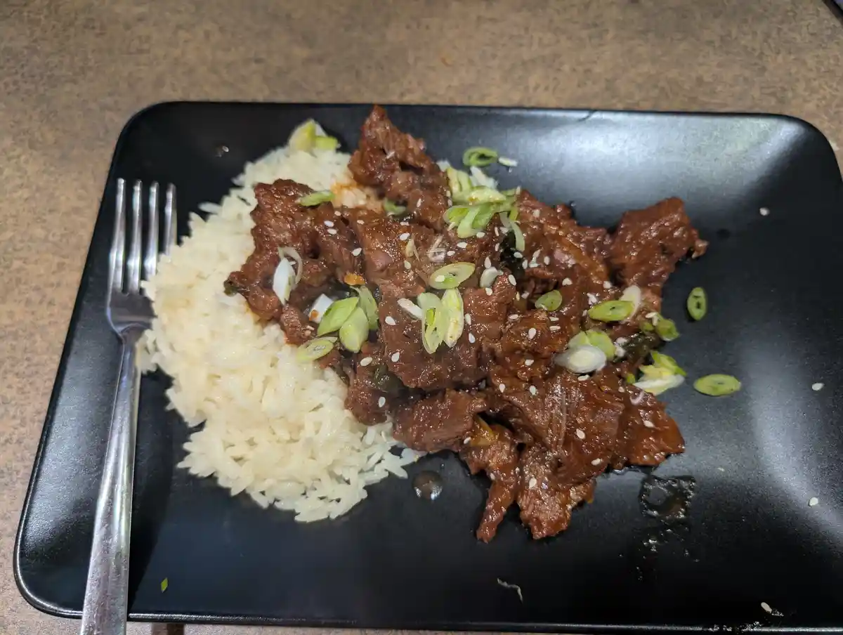 A black rectangular plate holds a serving of white rice and glossy brown meat, garnished with chopped green onions and white…