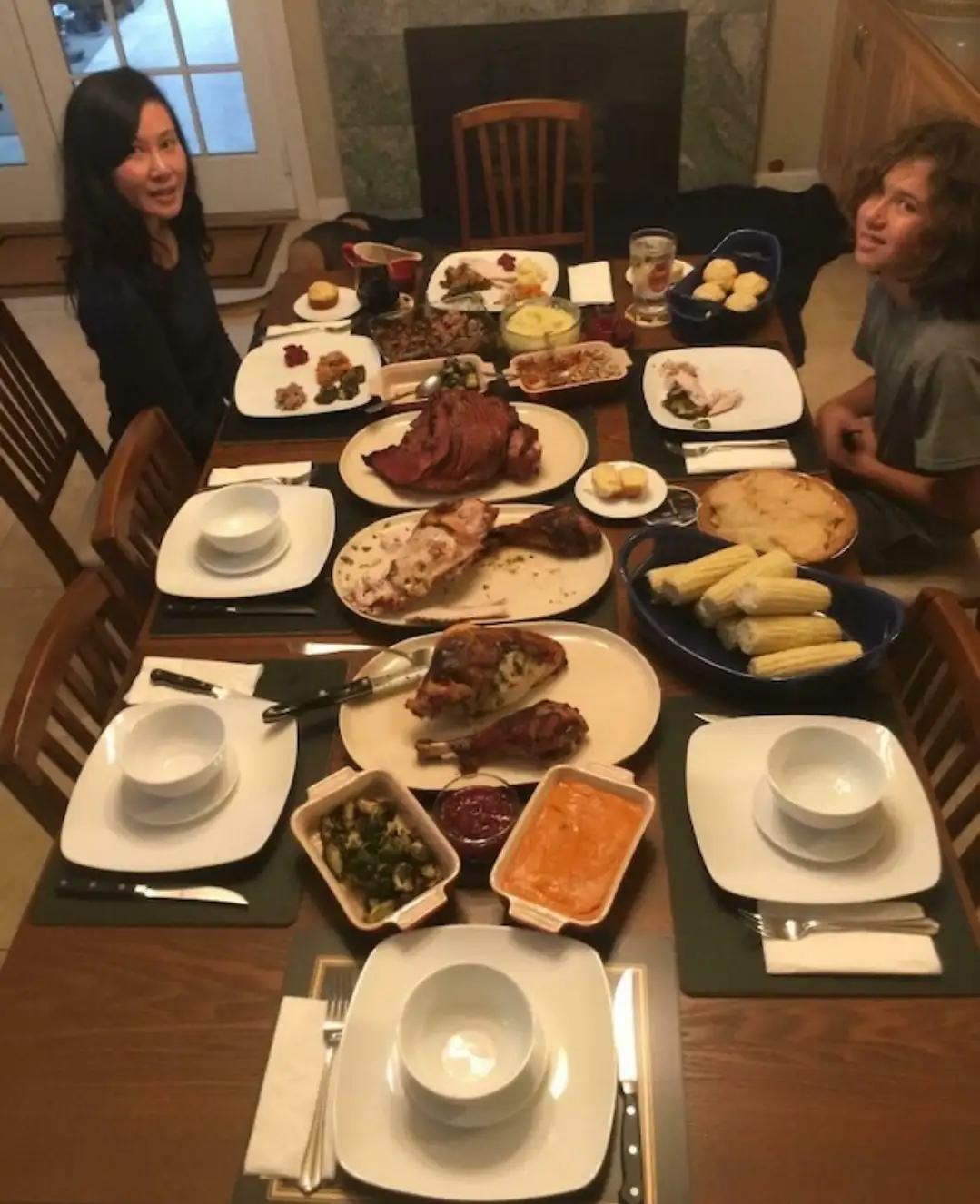A family dinner table is set with various dishes including ham, turkey, corn on the cob, and side dishes, with two people…