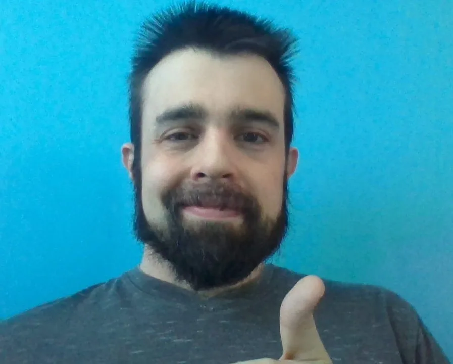 A man with a dark beard and short dark hair gives a thumbs-up gesture while smiling slightly against a solid blue…