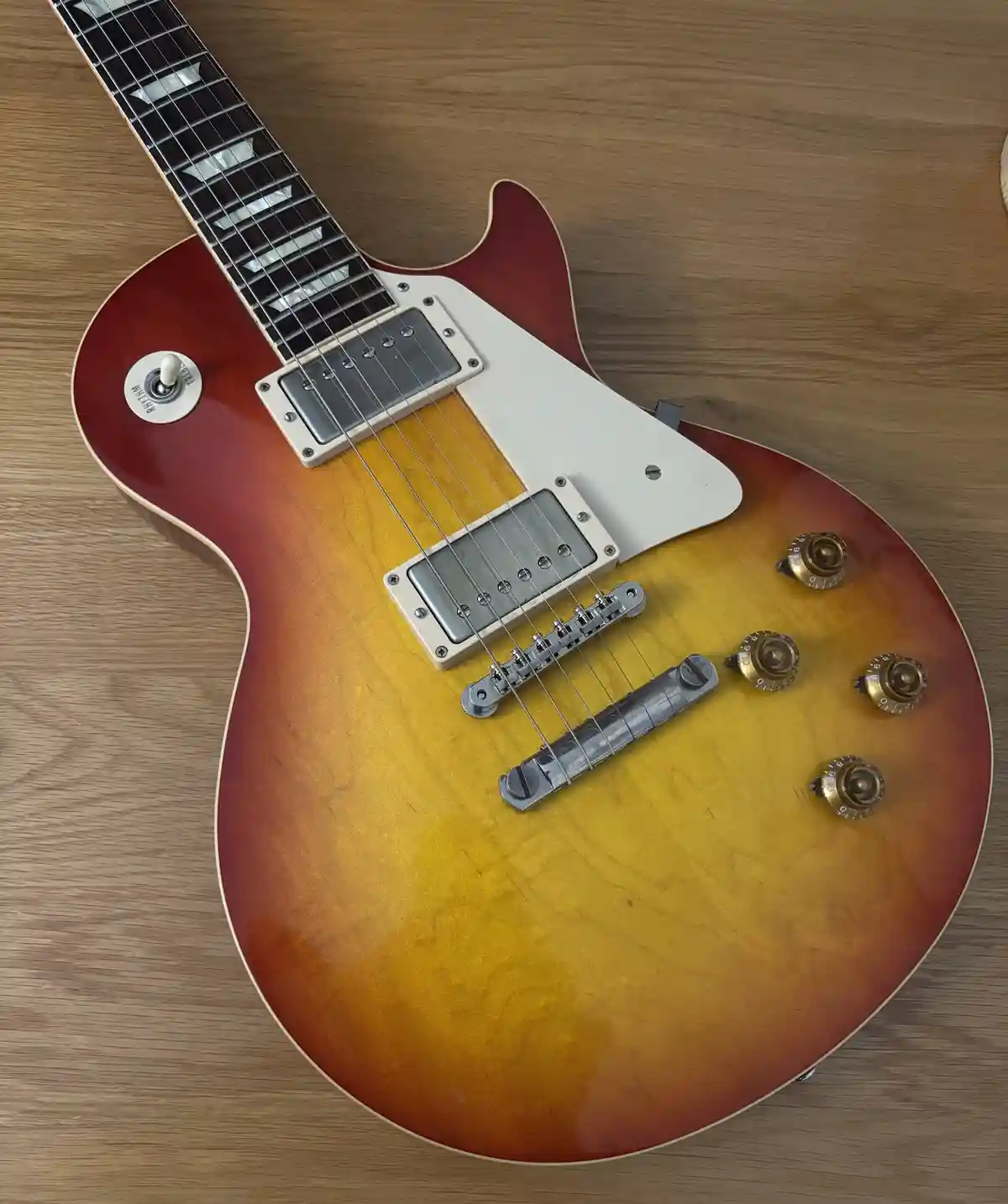 A sunburst-finish electric guitar with a white pickguard and gold knobs rests on a wooden surface. The overlaid text reads…