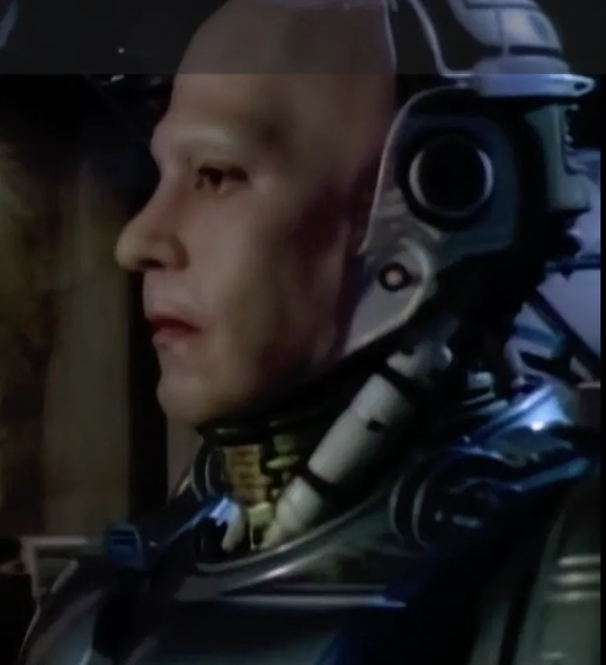 A close-up of a human face partially merged with robotic machinery, showing a human eye and cheek beside a mechanical neck…