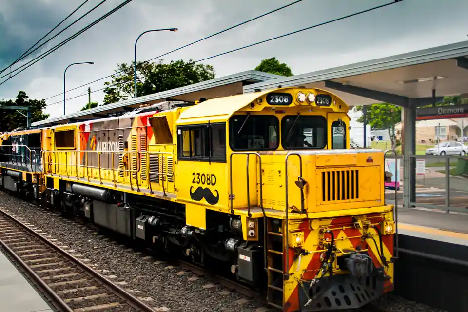 A yellow Aurizon locomotive, number 2308D, is pictured at the Dinmore train station. A large black mustache graphic has been…