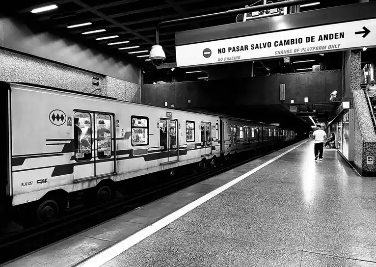 A black-and-white photo of a subway platform with a train stopped at the station. A sign above reads 'NO PASAR SALVO CAMBIO…