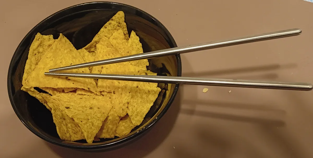 A black bowl filled with yellow, seasoned tortilla chips has a pair of metal chopsticks resting across the top. The image is…