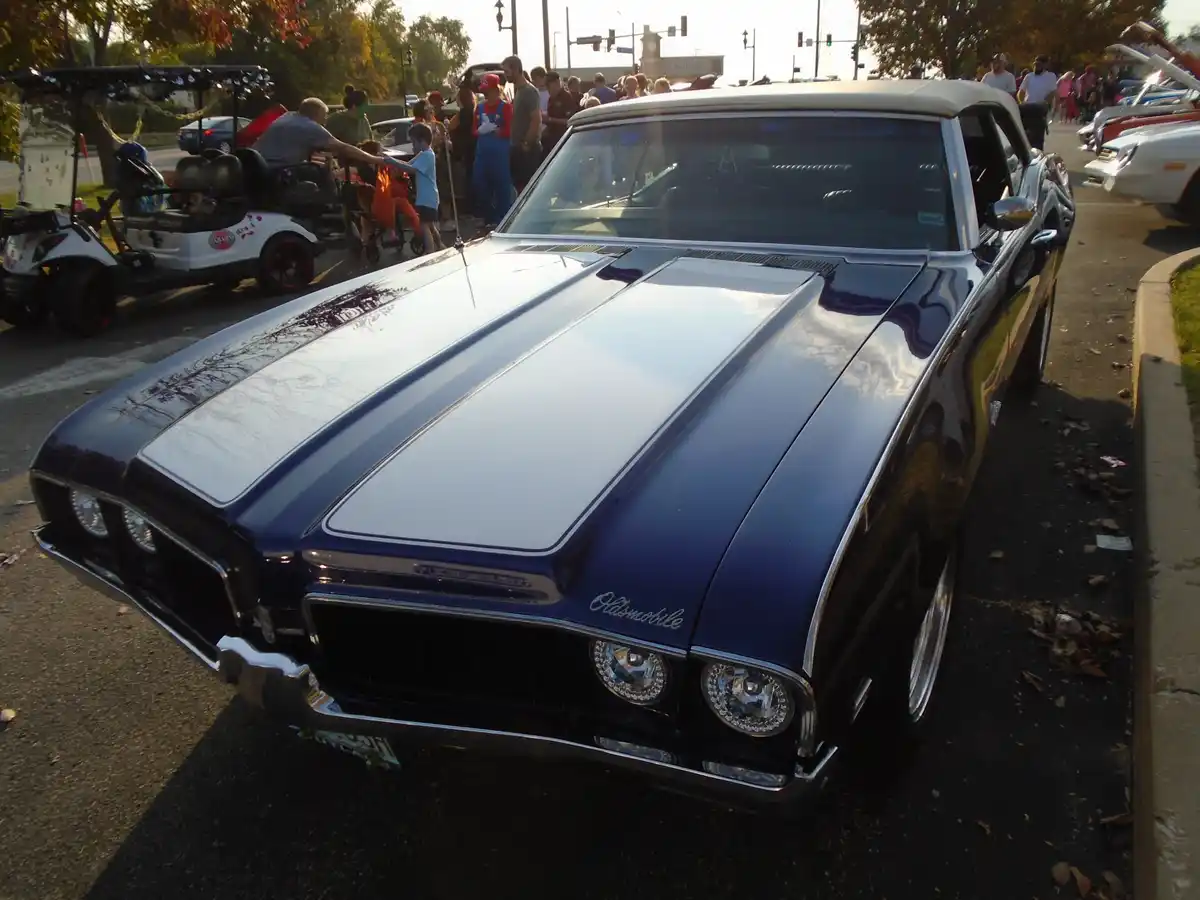 A shiny blue and white Oldsmobile classic car with chrome details is parked at an outdoor event, surrounded by people and…