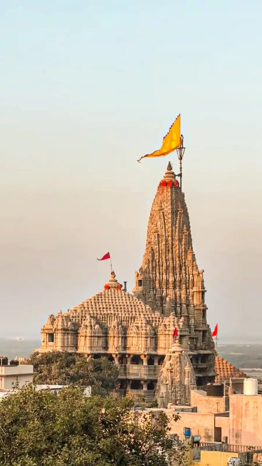 A tall, intricately carved stone temple with a large yellow flag at its peak and smaller red flags on other structures, set…