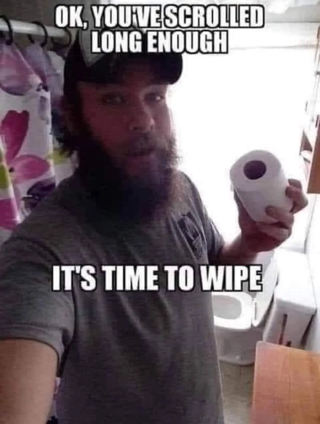A man with a beard and baseball cap stands in a bathroom, holding a roll of toilet paper. He looks directly at the camera…