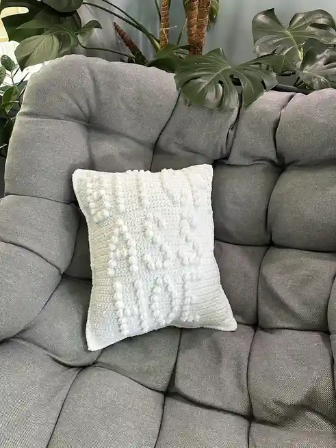 A white textured pillow with pom-pom details sits centered on a gray tufted couch, surrounded by large green monstera leaves…