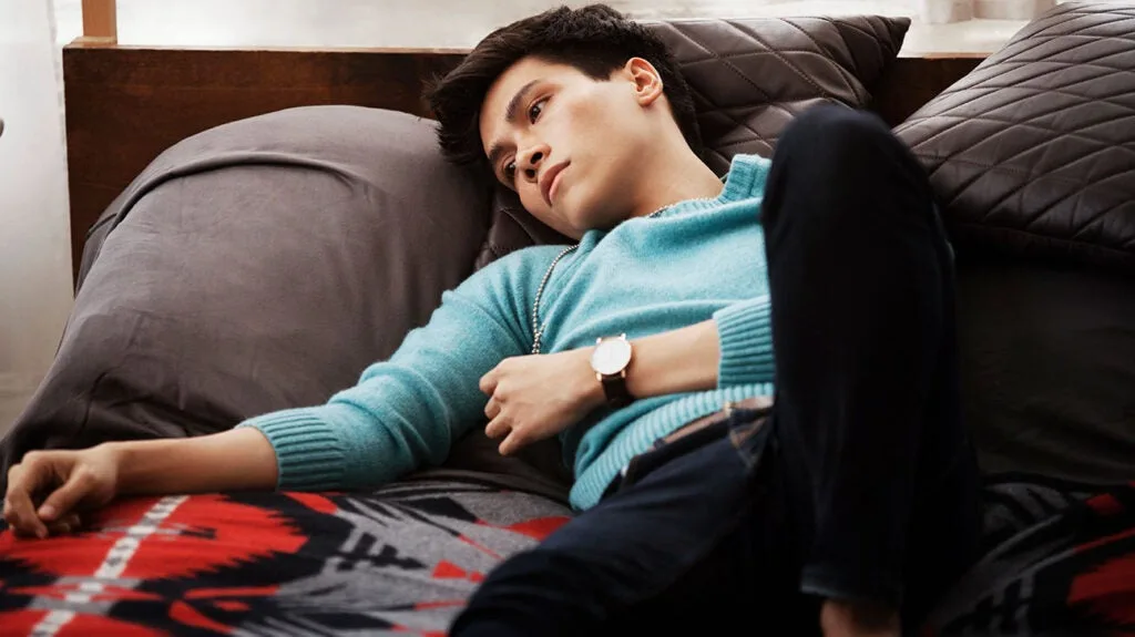a young man with dark hair lies on a bed with dark pillows and a red-and-gray patterned blanket, wearing a teal sweater and…