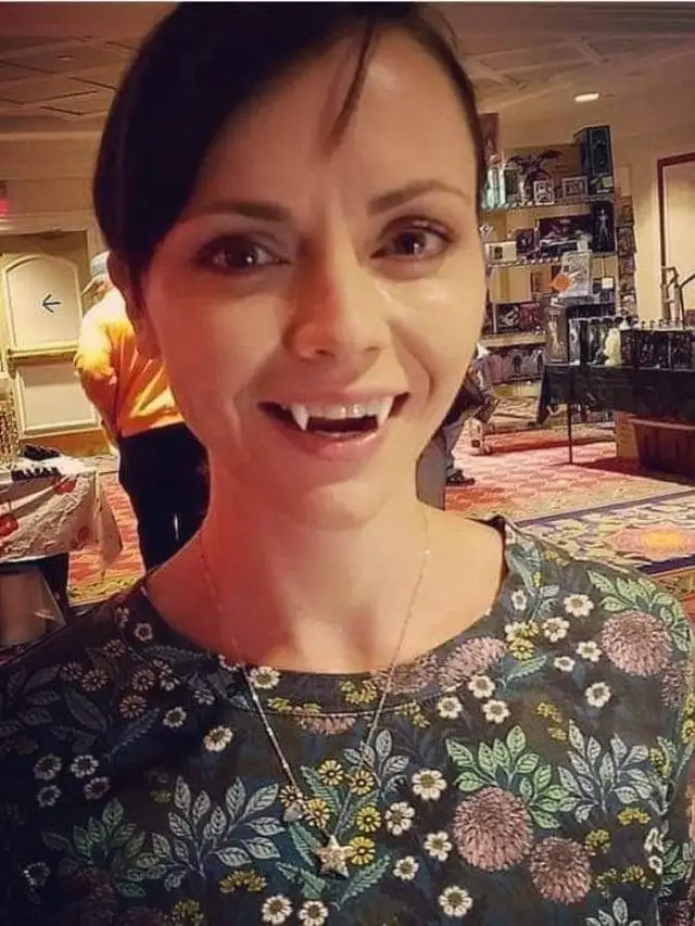 A woman with dark hair and a floral-patterned top smiles widely at the camera, revealing two prominent fangs. The background…
