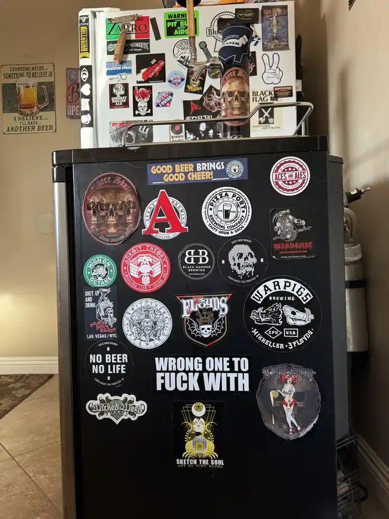 A black refrigerator covered in numerous beer-themed stickers from various breweries, including Three Skull, Avery Brewing…