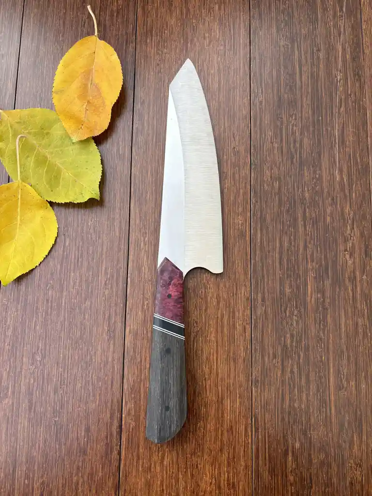 A chef's knife with a dark wooden handle and a red-brown patterned bolster lies on a dark brown wooden surface, next to…