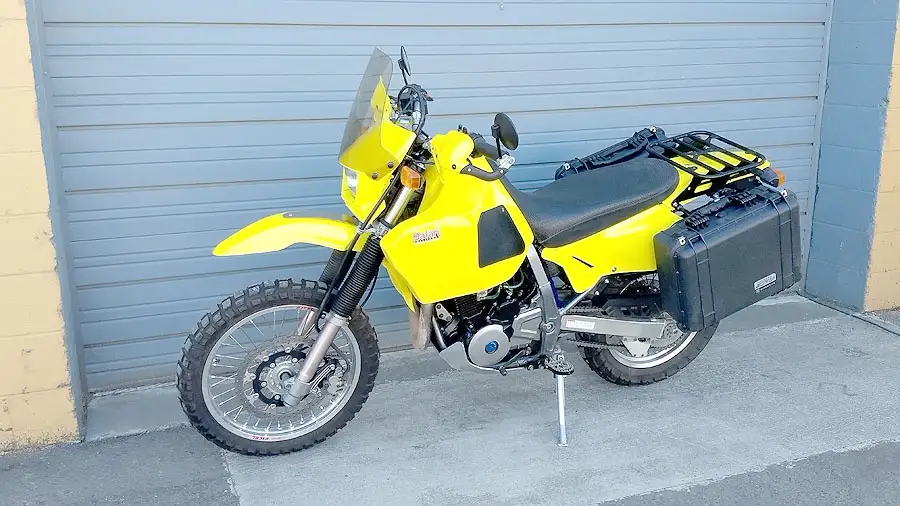 A bright yellow motorcycle, likely a Suzuki DR-Z, is parked on a concrete surface in front of a blue corrugated metal garage…