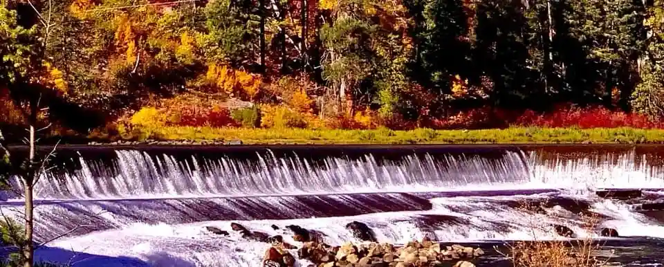 A scenic photo of a river flowing over a low dam, surrounded by vibrant autumn foliage in shades of orange, yellow, and…