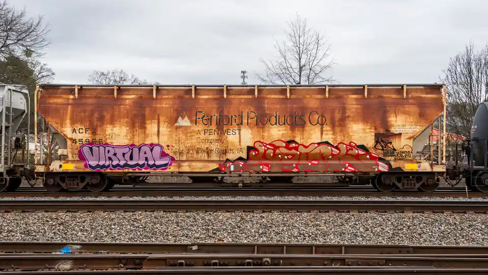 A rusty brown railroad hopper car with 'Peniford Products Co. A PENWEST Company Cedar Rapids, IA' printed on it, has the…