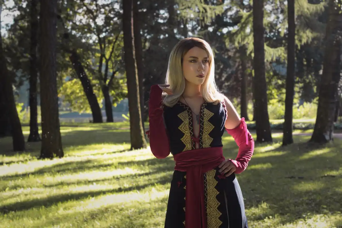 A person with blonde hair stands in a sunlit forest wearing a black and red outfit with gold embroidery, posing with one…