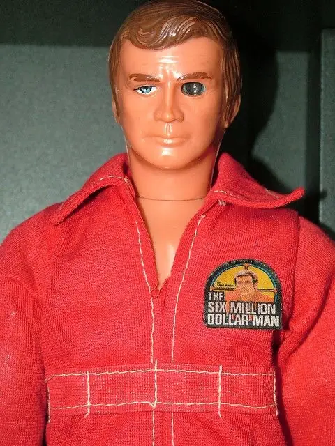 A close-up of a plastic action figure dressed in a red jumpsuit with white stitching, featuring a circular patch on the…