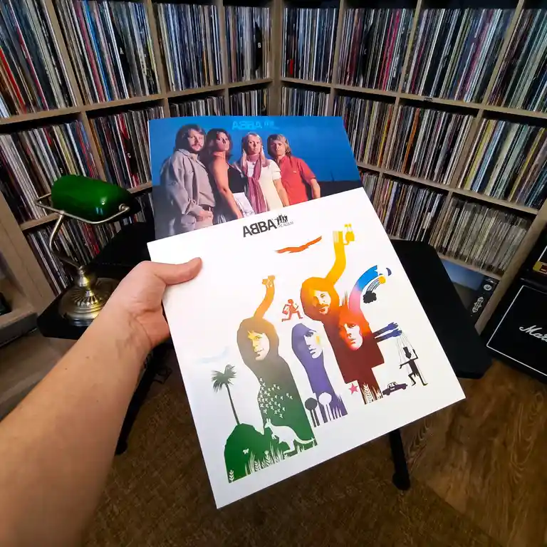 A hand holds up two ABBA vinyl record covers side-by-side: the original blue cover with the band's photo and the modern…