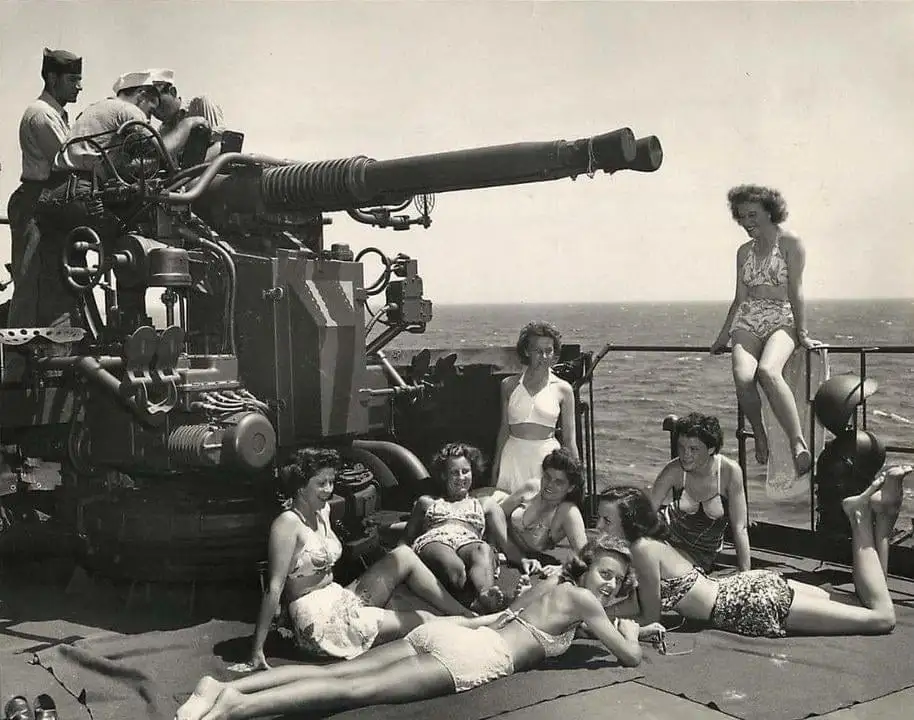 a vintage black-and-white photo shows several women in swimsuits lounging and sunbathing on the deck of a naval ship next to…