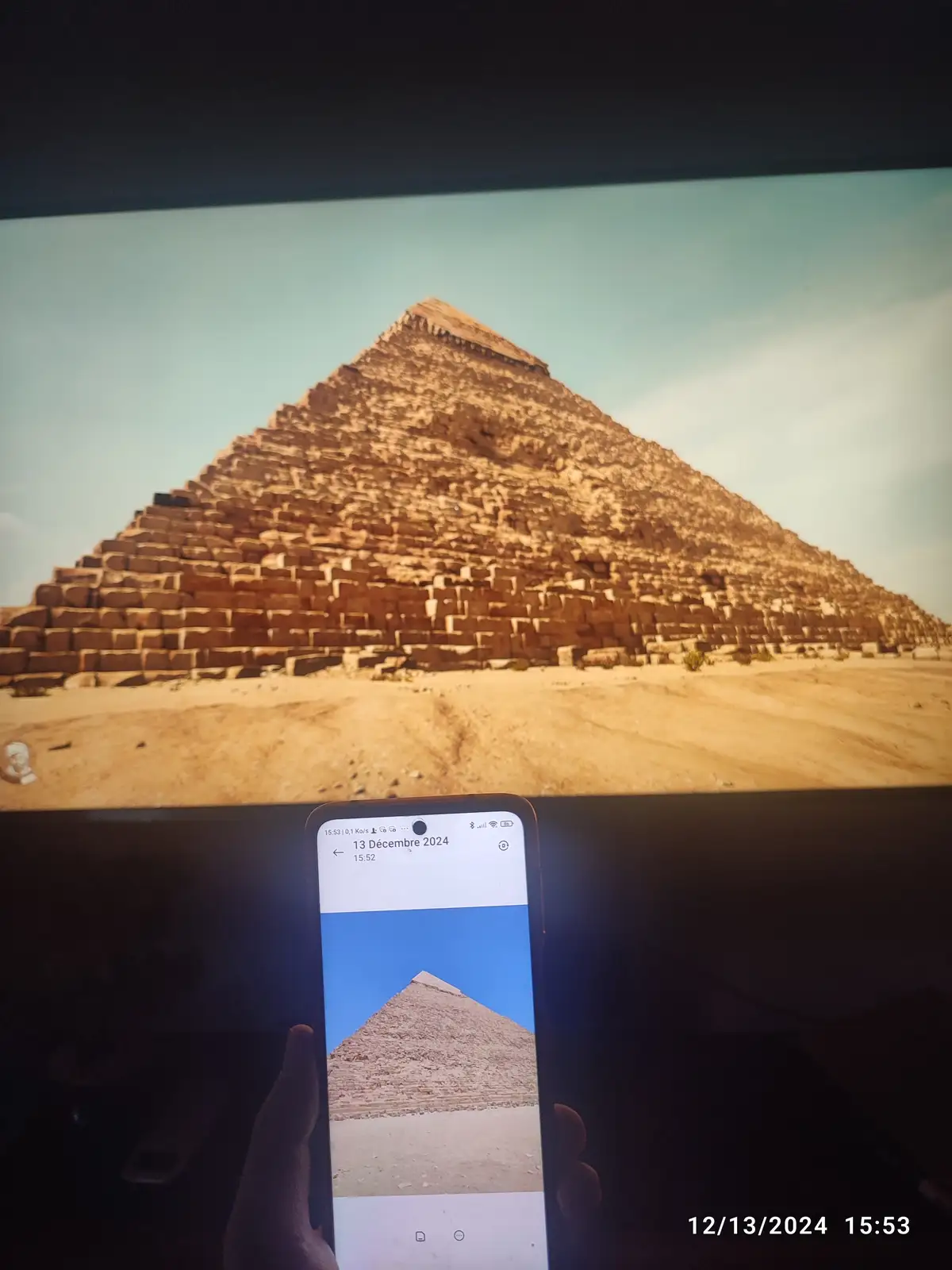A person holds a smartphone displaying a photo of a pyramid, while a larger screen behind them shows the same pyramid but…