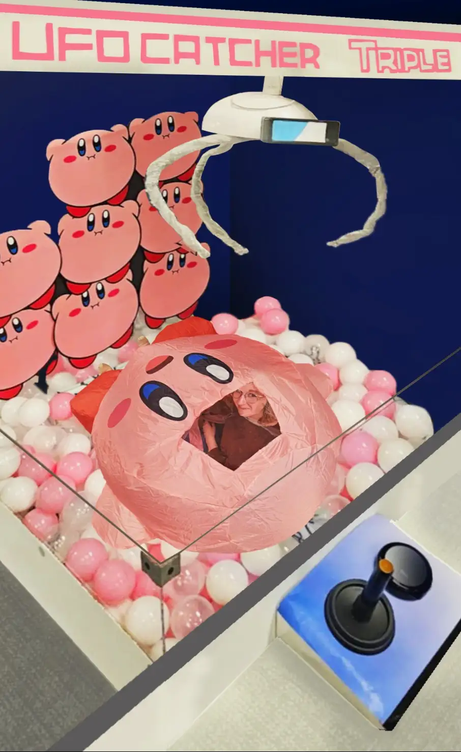 A person with glasses is visible inside a large pink Kirby plush toy, which is situated inside a claw machine filled with…