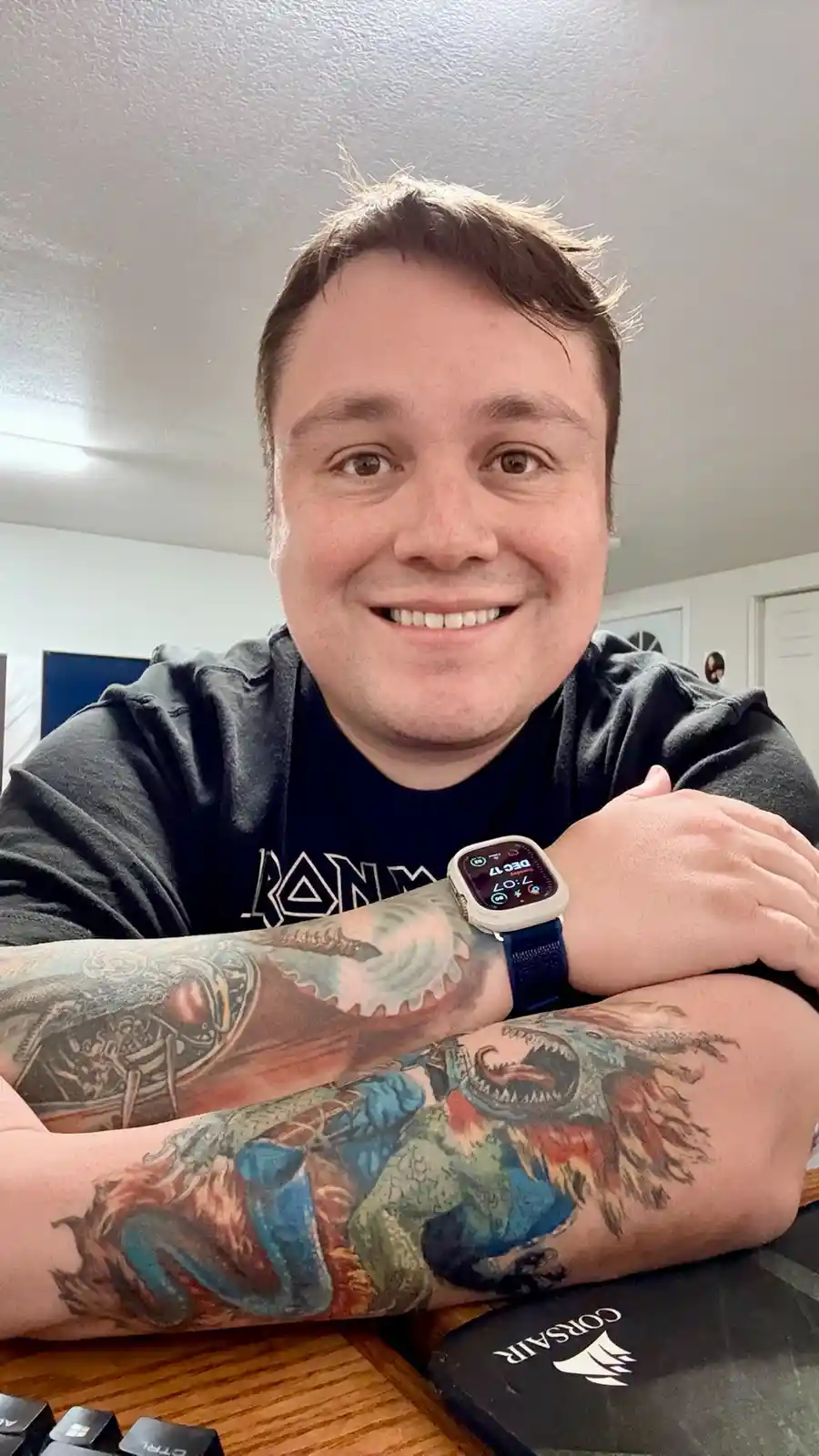 A smiling man with heavily tattooed forearms rests his arms on a desk, wearing a smartwatch showing the time as 7:07. His…