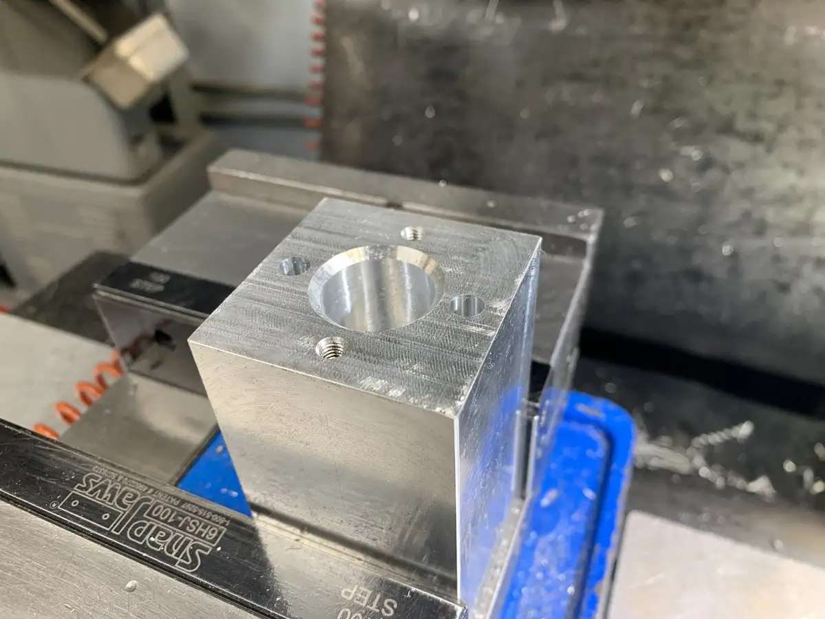 A close-up photo shows a freshly machined metal block with a central cylindrical hole and four smaller holes around it…