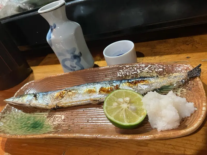 A grilled whole fish, likely a Japanese mackerel or similar, rests on a textured ceramic plate alongside a lime wedge and a…