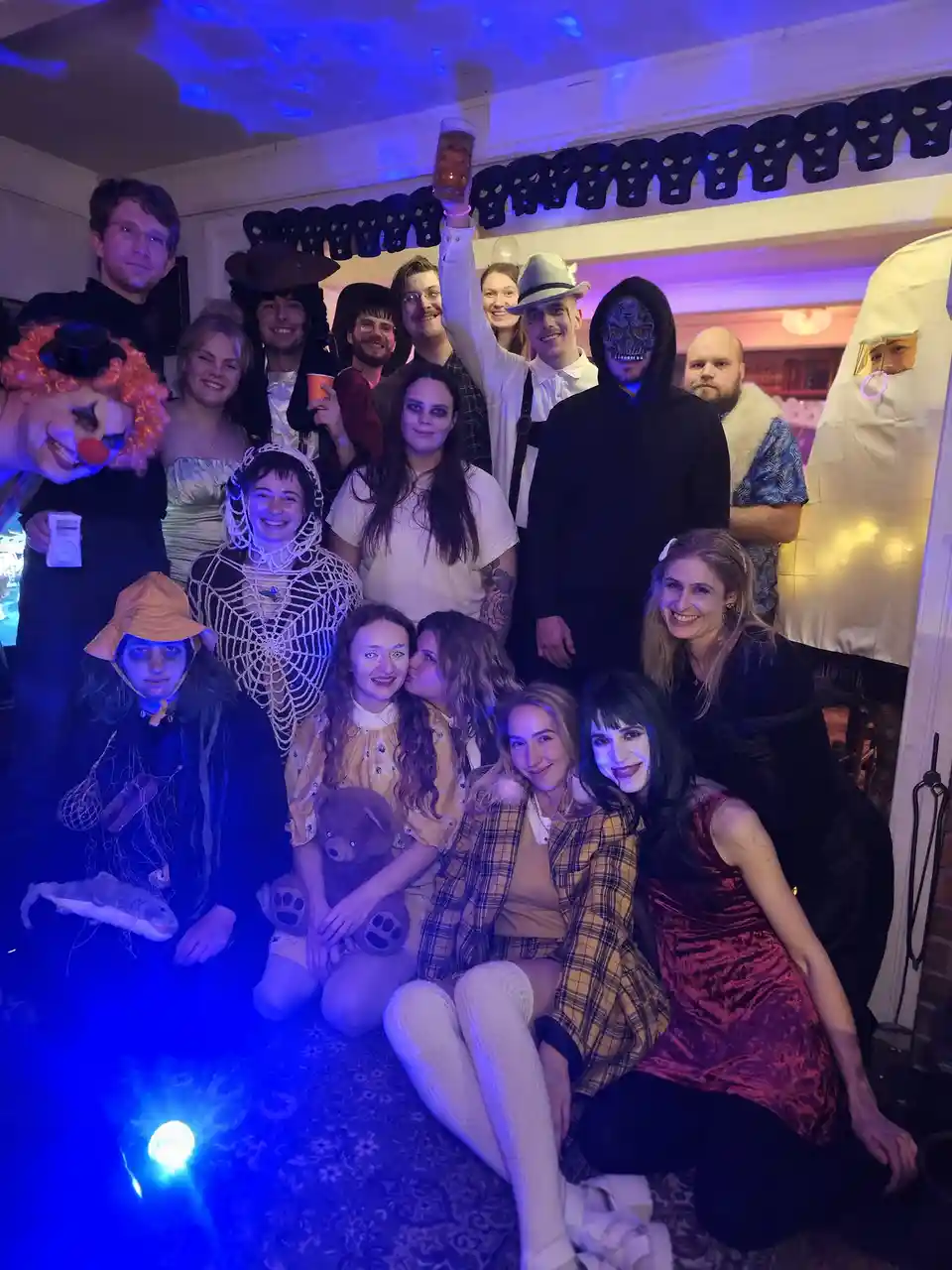 A group of people dressed in various Halloween costumes including a clown, spiderweb outfit, ghost, skull mask, and others…