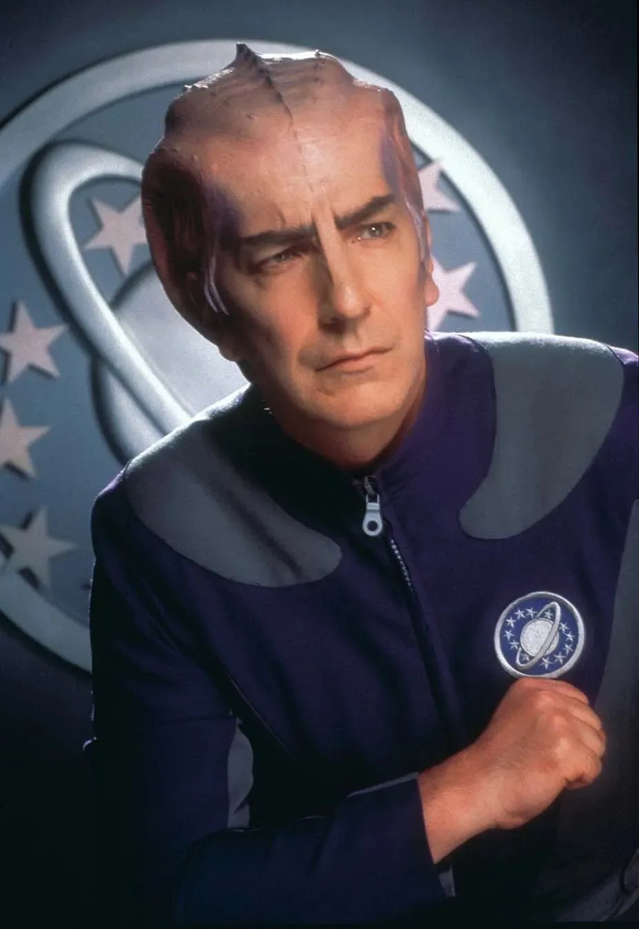 A man with a bald, scarred head and a serious expression wears a dark blue uniform with a circular emblem featuring a planet…