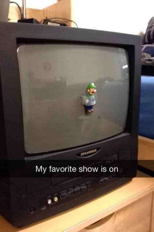 A small Luigi figurine is stuck to the dusty screen of an old black Sylvania CRT television, which sits on a wooden surface…