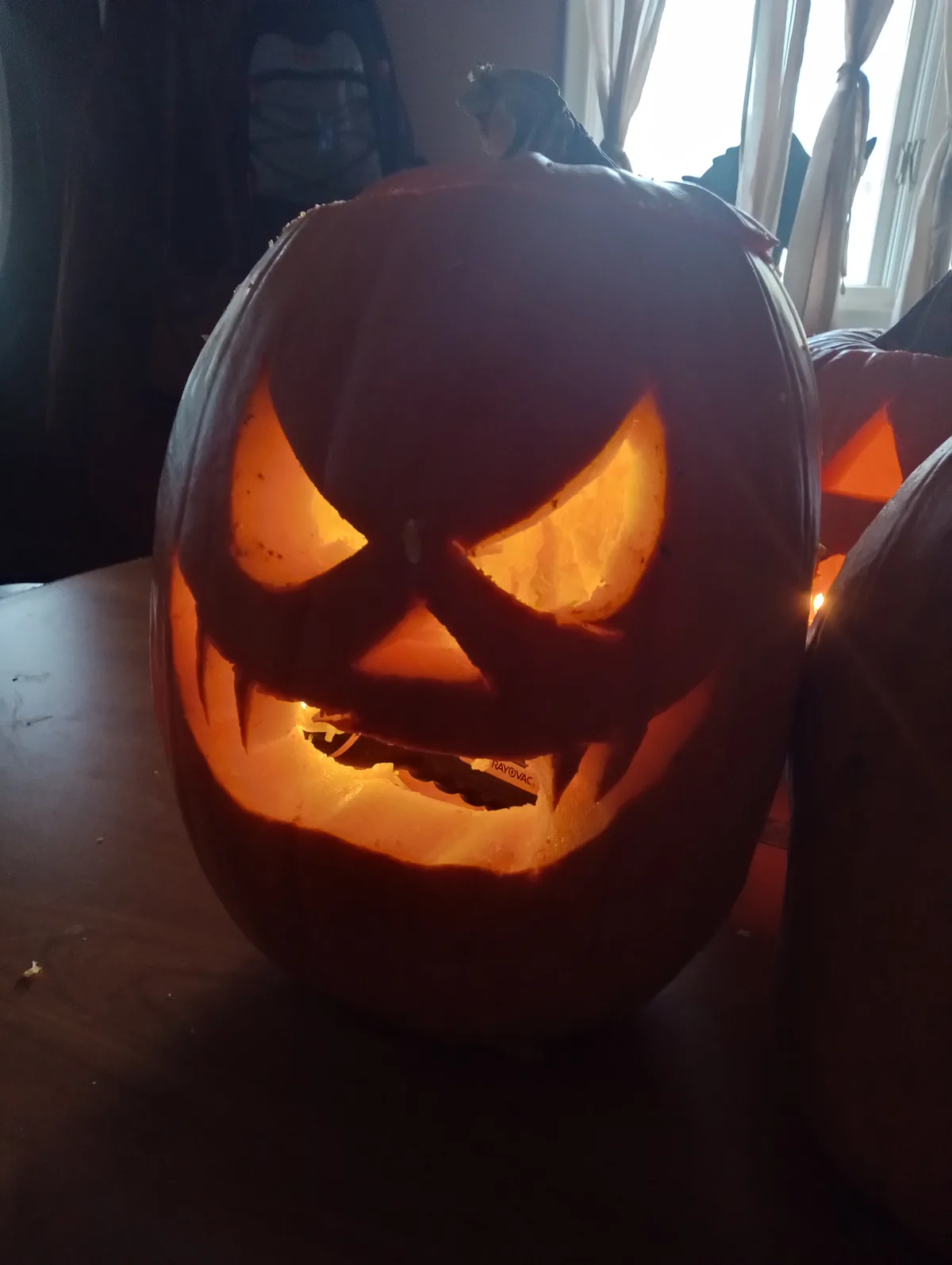 A close-up of a carved Halloween pumpkin with glowing eyes and a jagged mouth, lit from within by a battery visible inside…