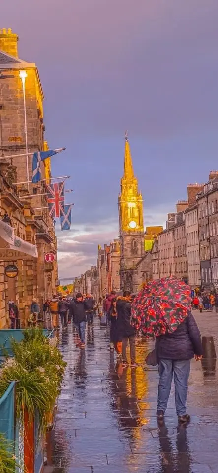 A person walks down a wet, cobblestone street in Edinburgh, holding a large, colorful floral-patterned umbrella. The street…