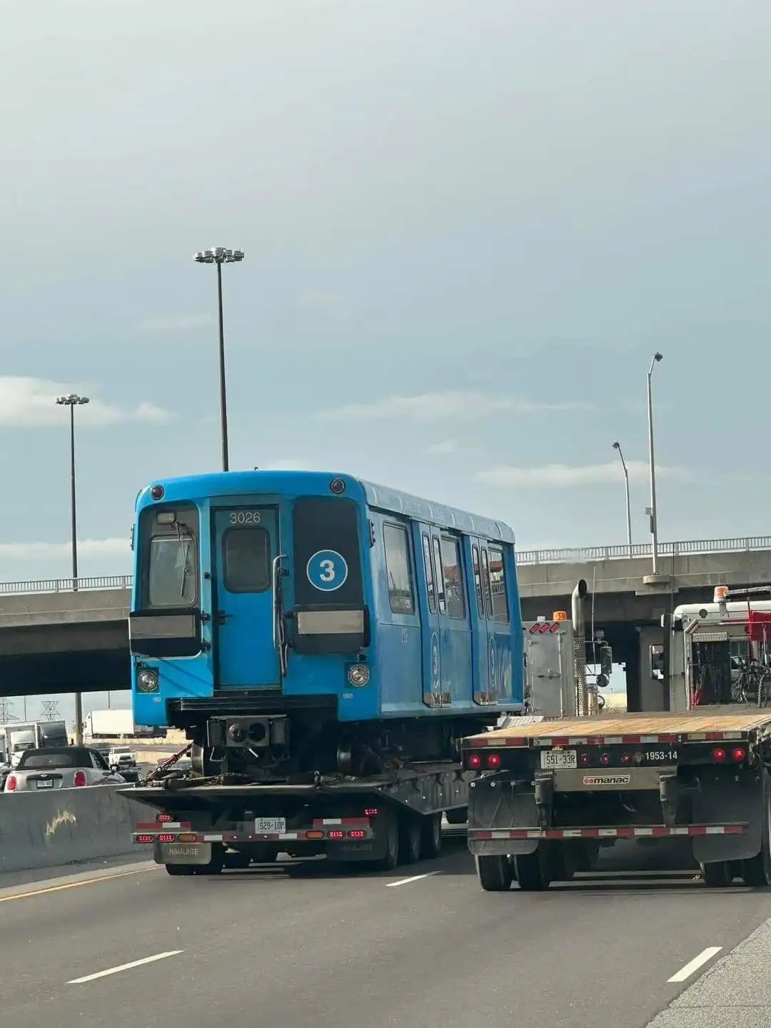 A blue New York City subway car, marked with the number 3, is being transported on a flatbed truck while driving on a…