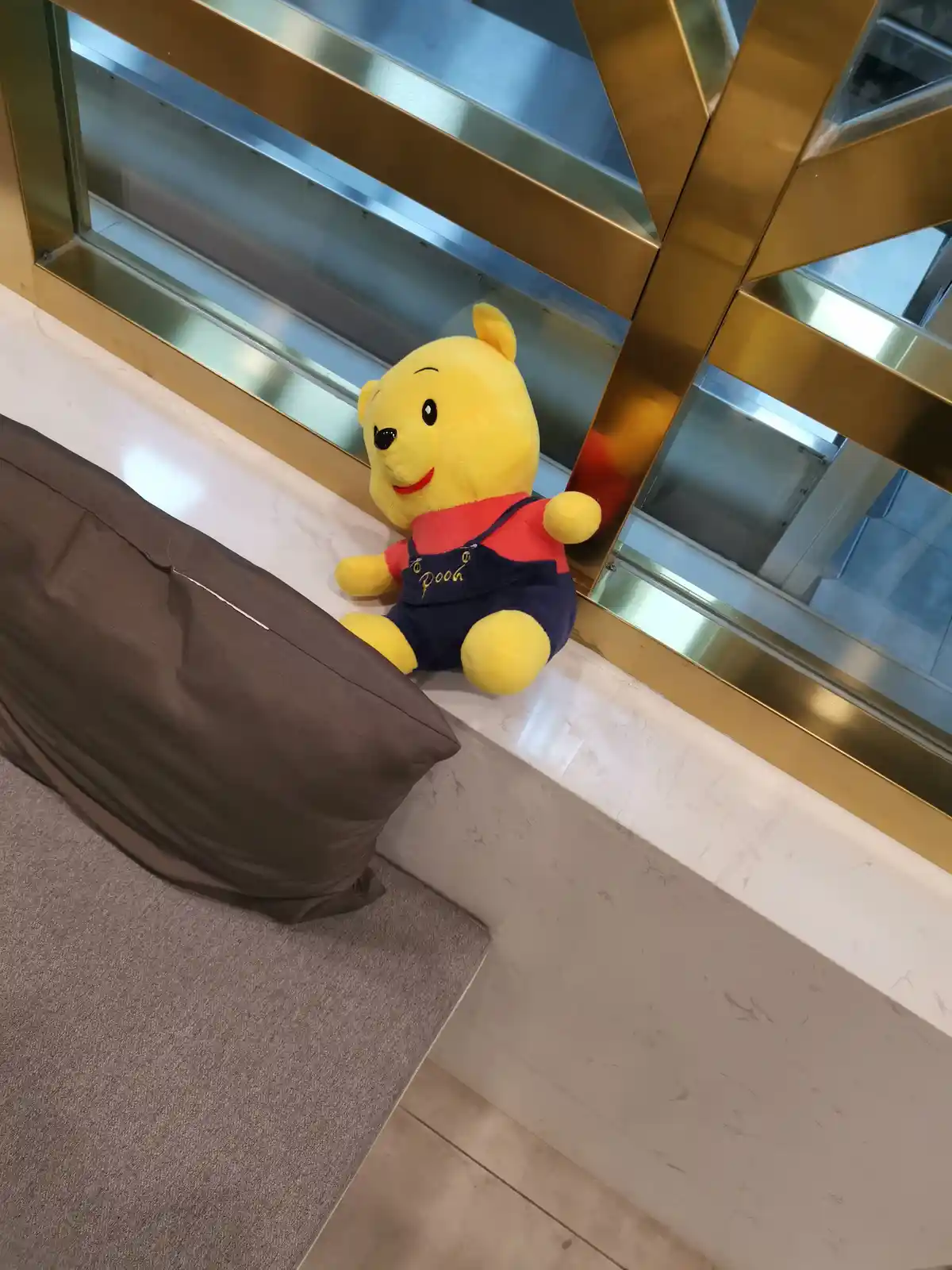 A yellow plush Winnie the Pooh doll wearing a red shirt and blue overalls sits on a white marble ledge next to a brown…