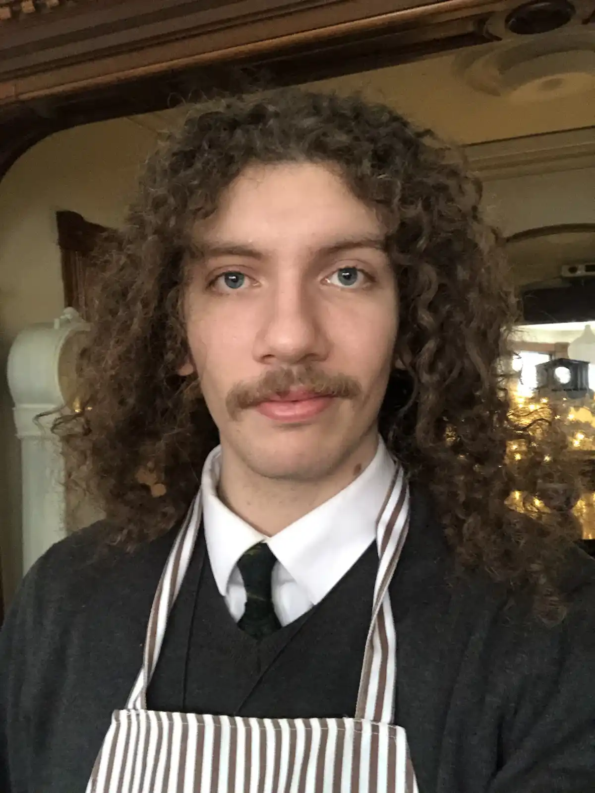 A young man with long curly brown hair and a mustache looks directly at the camera, wearing a white collared shirt, dark…