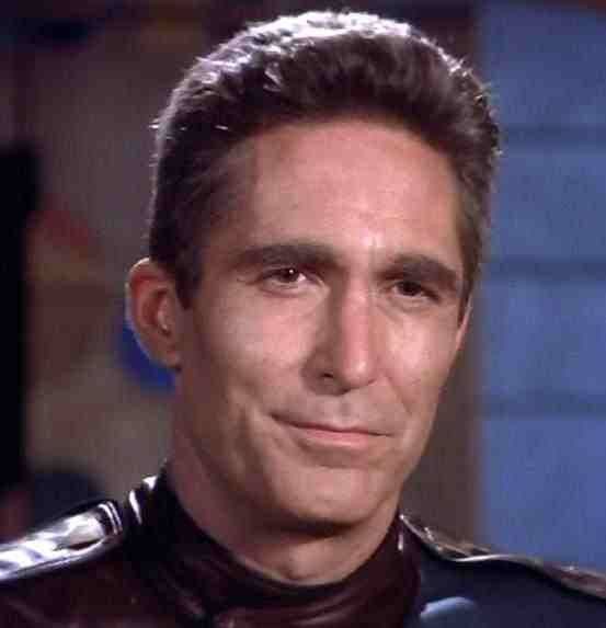 A close-up of a man with short dark hair and a subtle smirk, wearing a dark uniform with shoulder epaulets, looking directly…