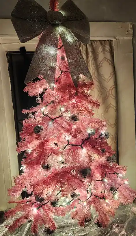 A brightly lit pink artificial Christmas tree decorated with disco ball ornaments and string lights, topped with a large…