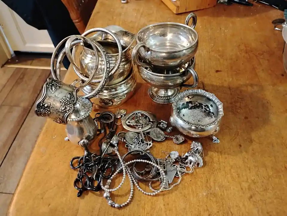 A wooden table is covered with a large collection of tarnished silver items, including stacked bowls, pitchers with ornate…