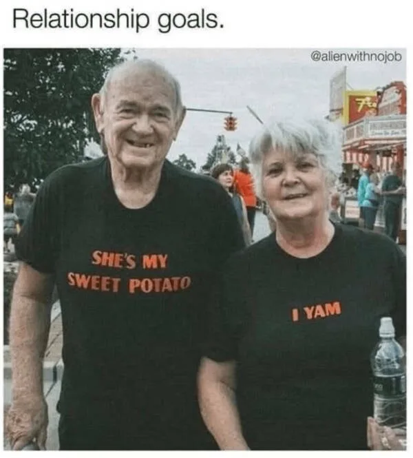 An elderly couple smiles at the camera while standing outdoors at what appears to be a fair or festival. The man wears a…