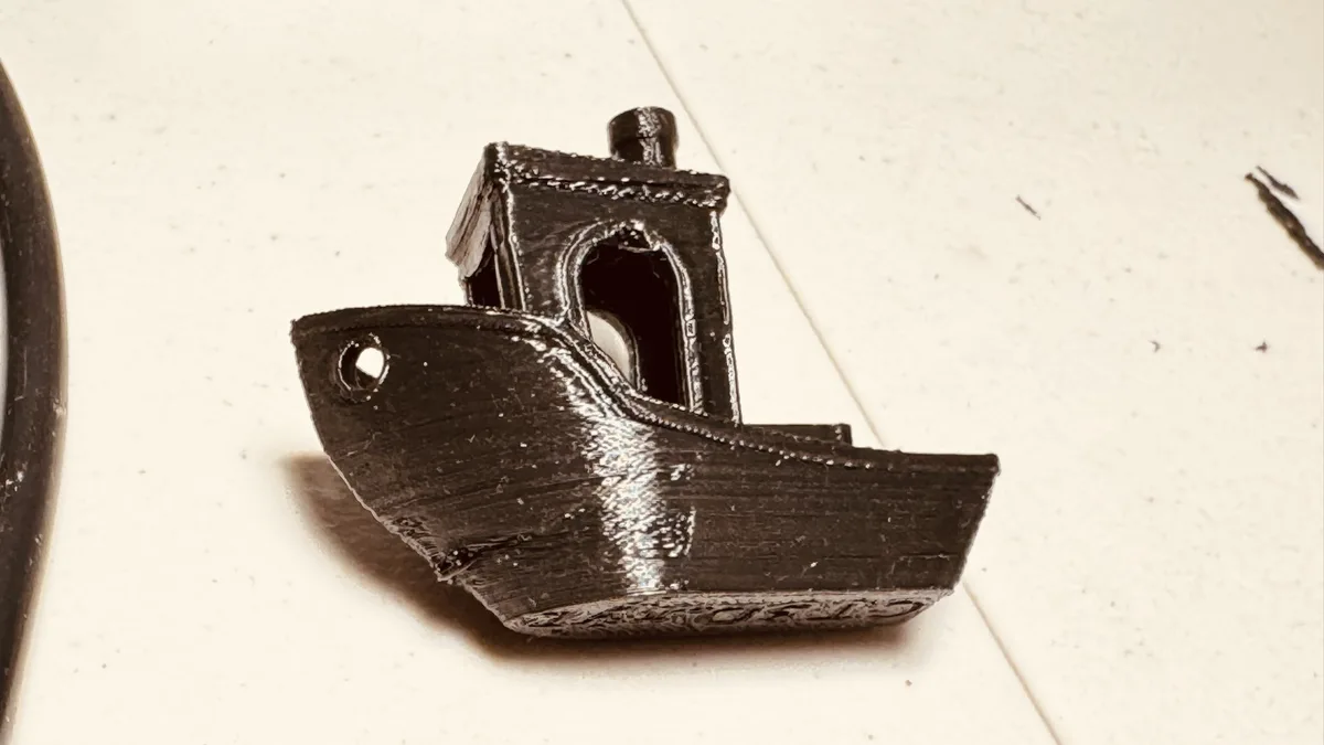 A small, black 3D-printed model of a boat with visible layer lines and a crude cabin sits on a light-colored tiled surface…