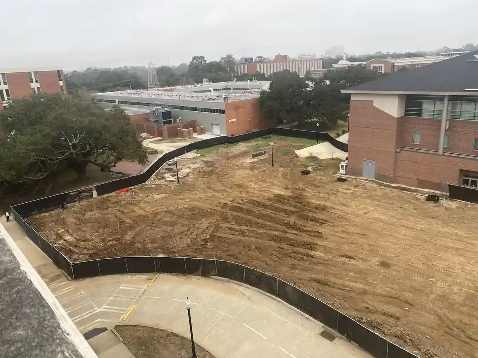 An aerial view of a large, muddy construction site surrounded by black fencing, with brick buildings and trees in the…