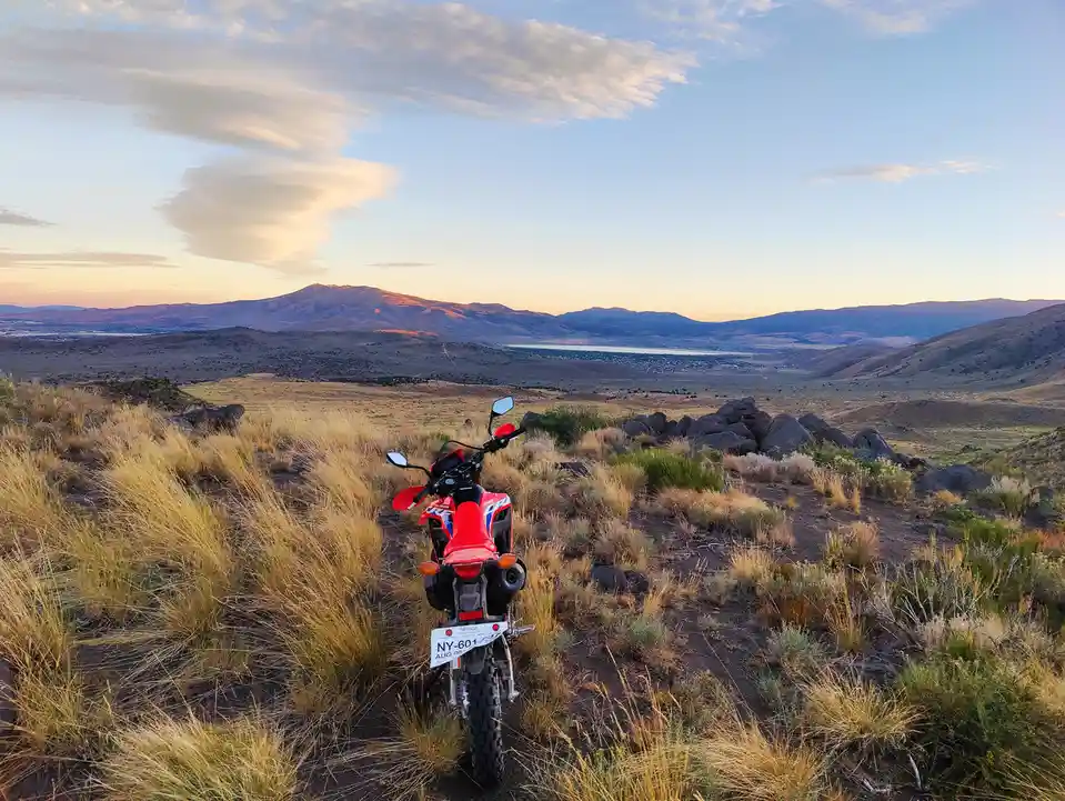 A red dirt bike is parked on a grassy hill overlooking a vast desert valley with distant mountains and a lake at sunset. The…