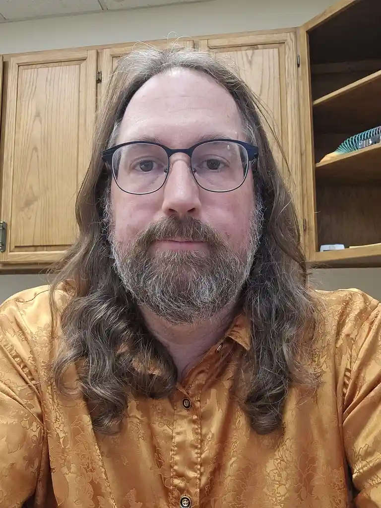 A man with long brown and gray hair, a beard, and black-rimmed glasses stares directly at the camera while wearing a shiny…
